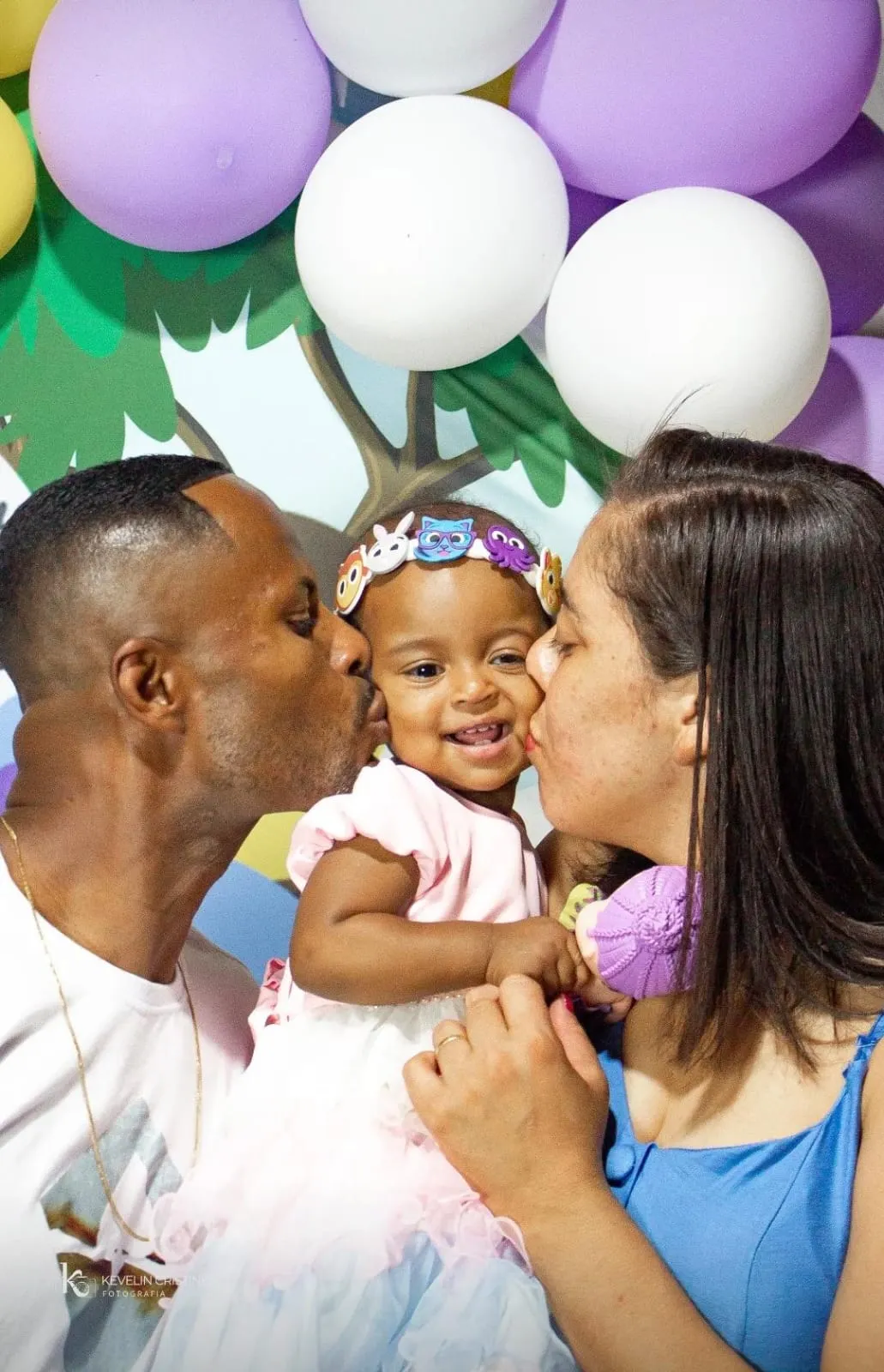 Pai e mãe beijando o rosto da filha sorridente, em festa de aniversário com balões lilás e brancos