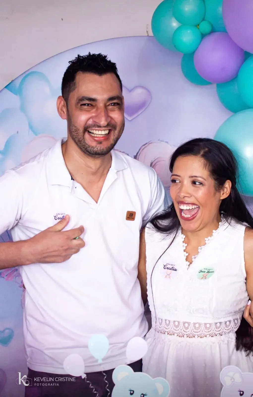 Casal sorrindo em frente ao painel de balões verde-água e lilás de um chá revelação