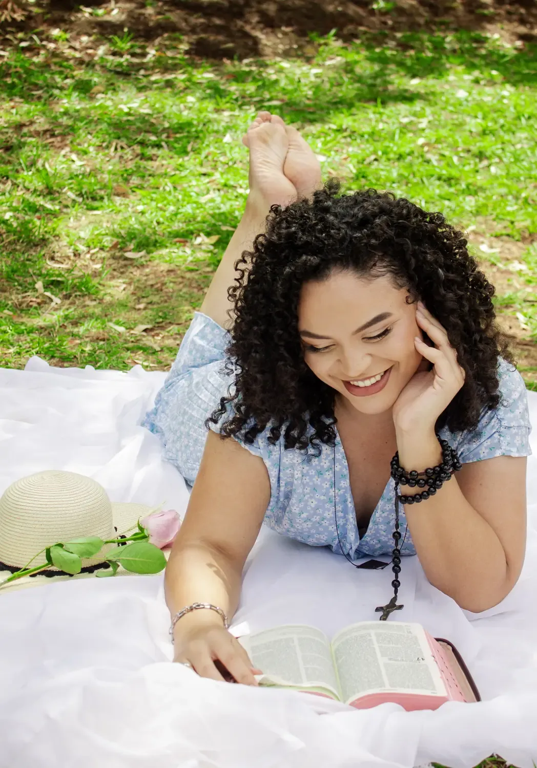 Mulher sorrindo deitada na grama, terço e livro à frente