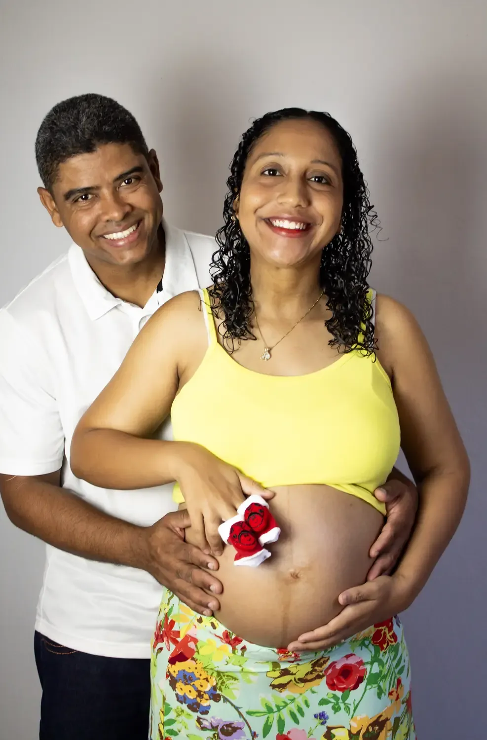 Casal com as mãos na barriga de grávida, segurando sapatinhos vermelhos de bebê
